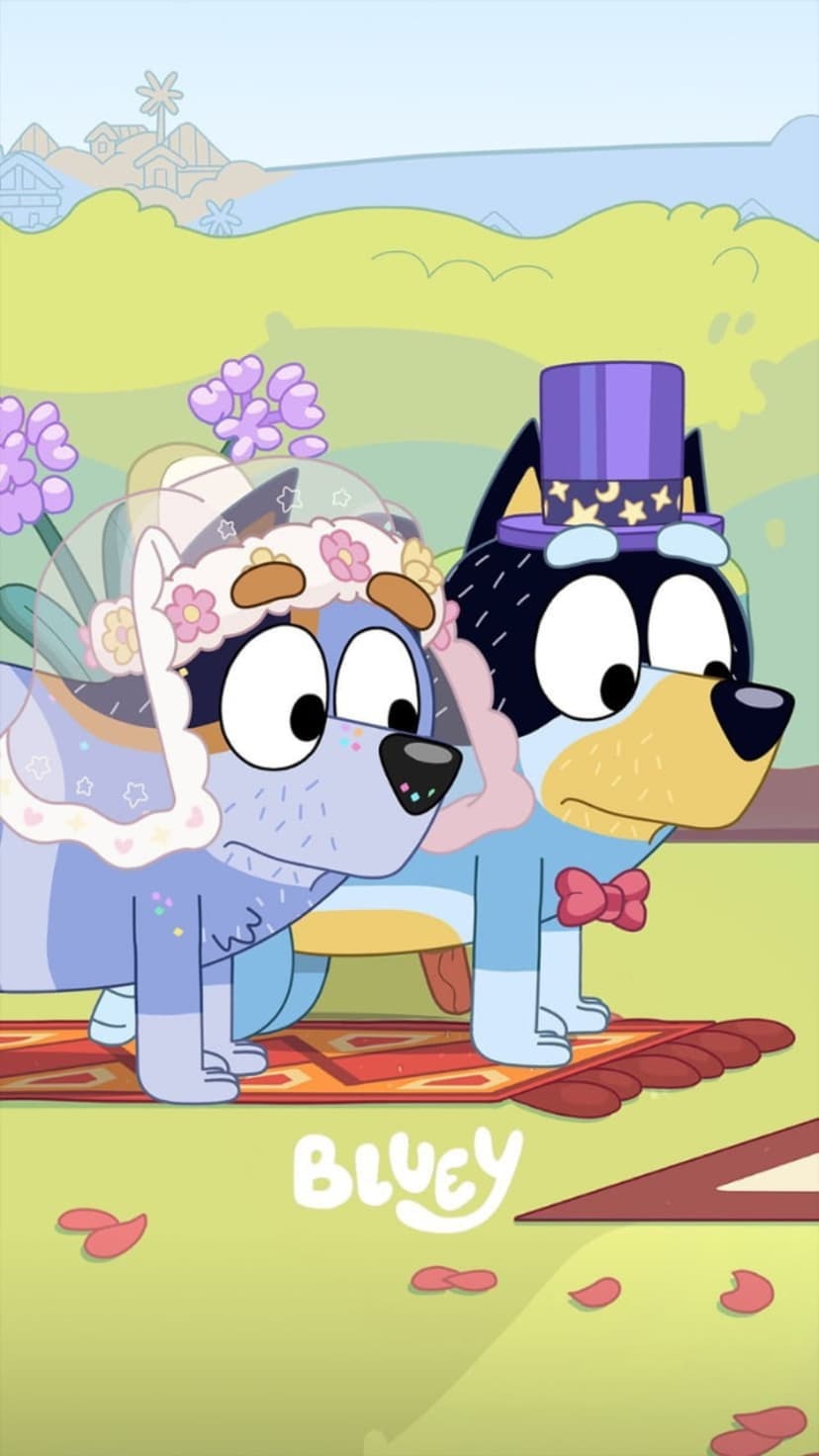 Bluey wearing a floral veil and Bingo in a purple top hat stand on a rug with rose petals under a blue sky.