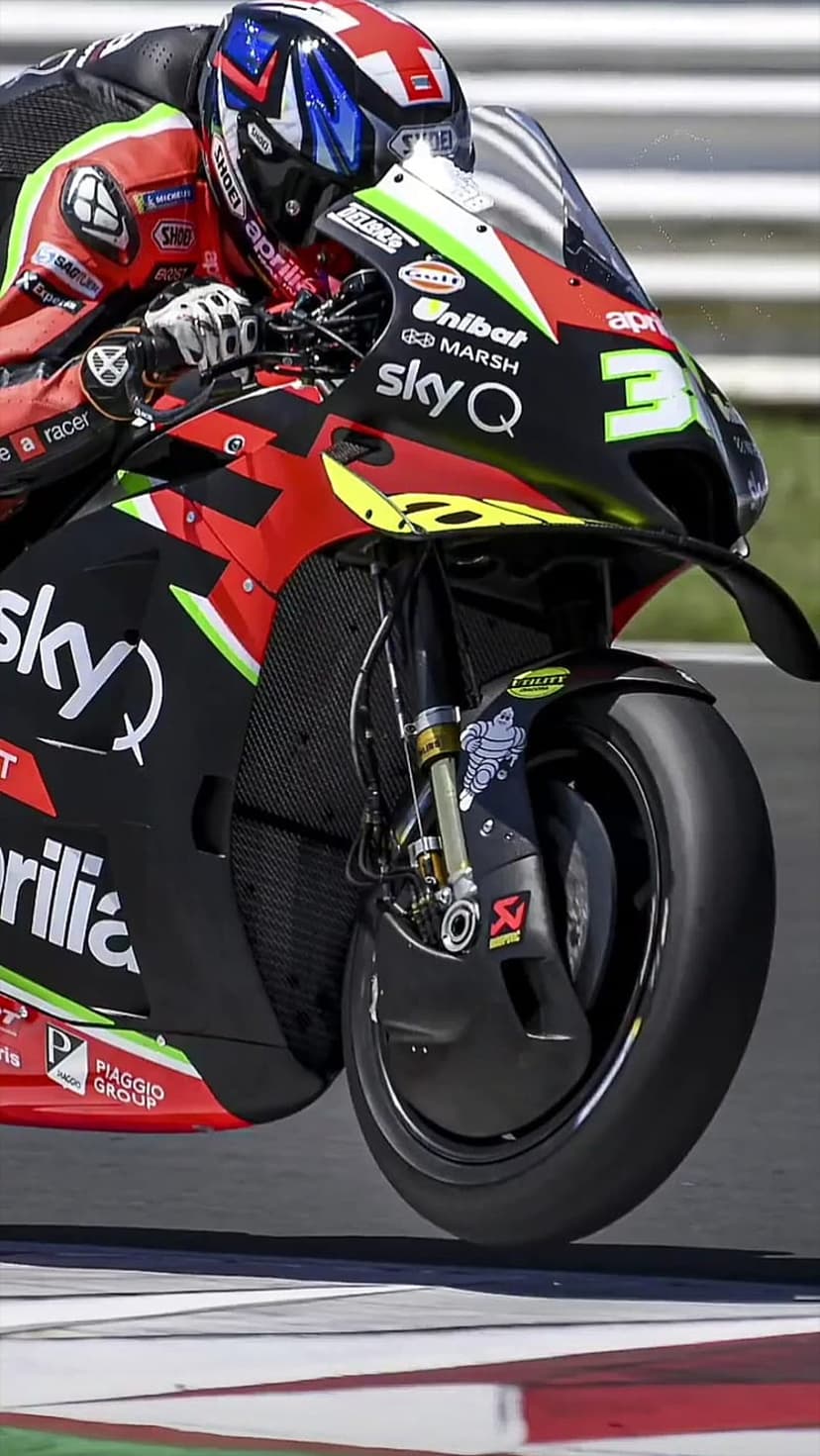 Aprilia MotoGP rider leans low into a racetrack corner, wearing a red and white helmet on a colorful racing bike.