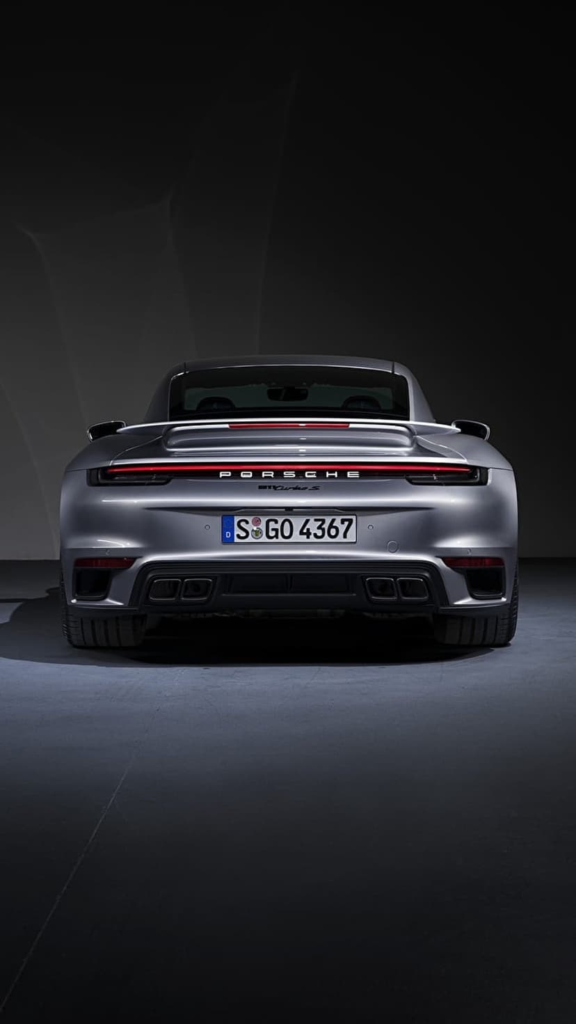 Porsche 911 Turbo S rear view with a grey exterior, glowing red taillights, and quad exhausts in a dark studio.