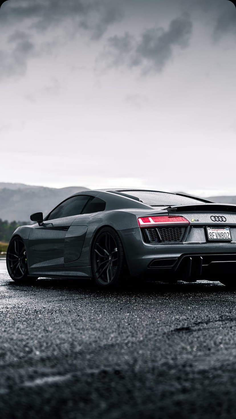 Audi R8 rear view with glowing red taillights reflecting on a wet asphalt road against misty mountain peaks.