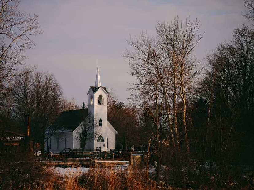 White church steeple rises behind dark, leafless tree branches on a serene and snow-dusted winter landscape.