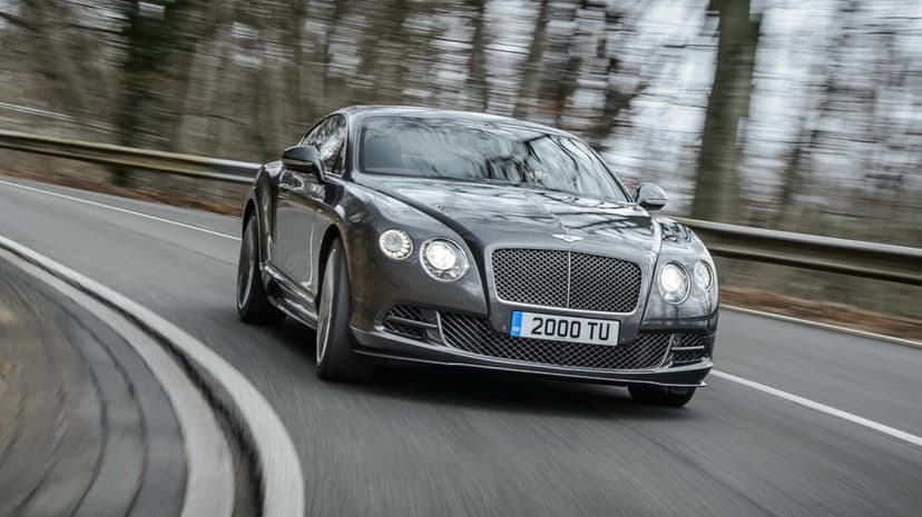 Dark grey Bentley luxury car driving fast on a curved forest road with motion blur and glowing headlights.