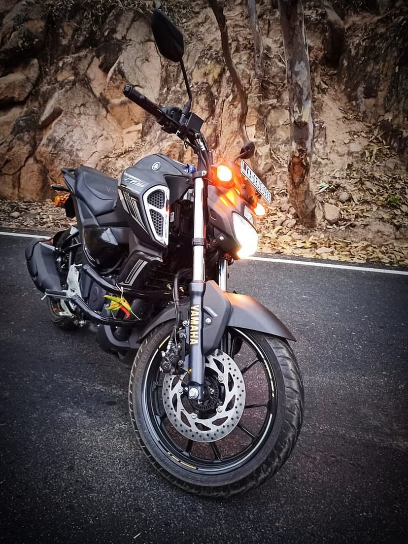 Black Yamaha motorcycle with glowing headlights parked on a paved road beside a steep, rocky hillside at twilight.