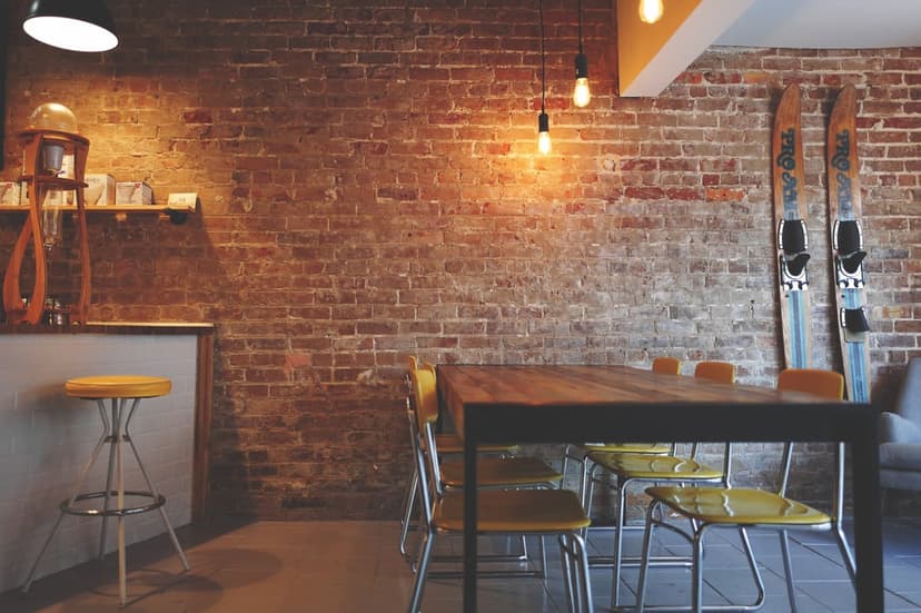Weathered brick wall behind a wooden table with yellow chairs and vintage skis under warm pendant lighting.