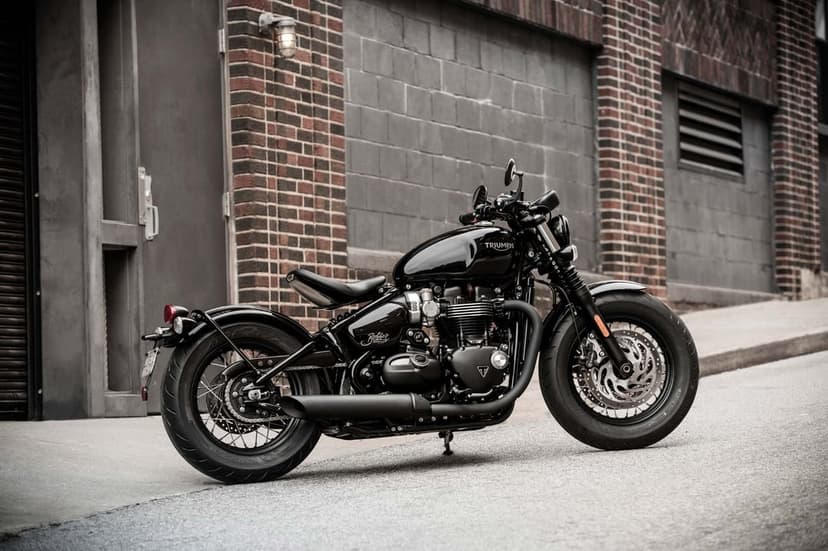 Black Triumph Bonneville Bobber motorcycle parked on a street against a grey cinder block and red brick wall.