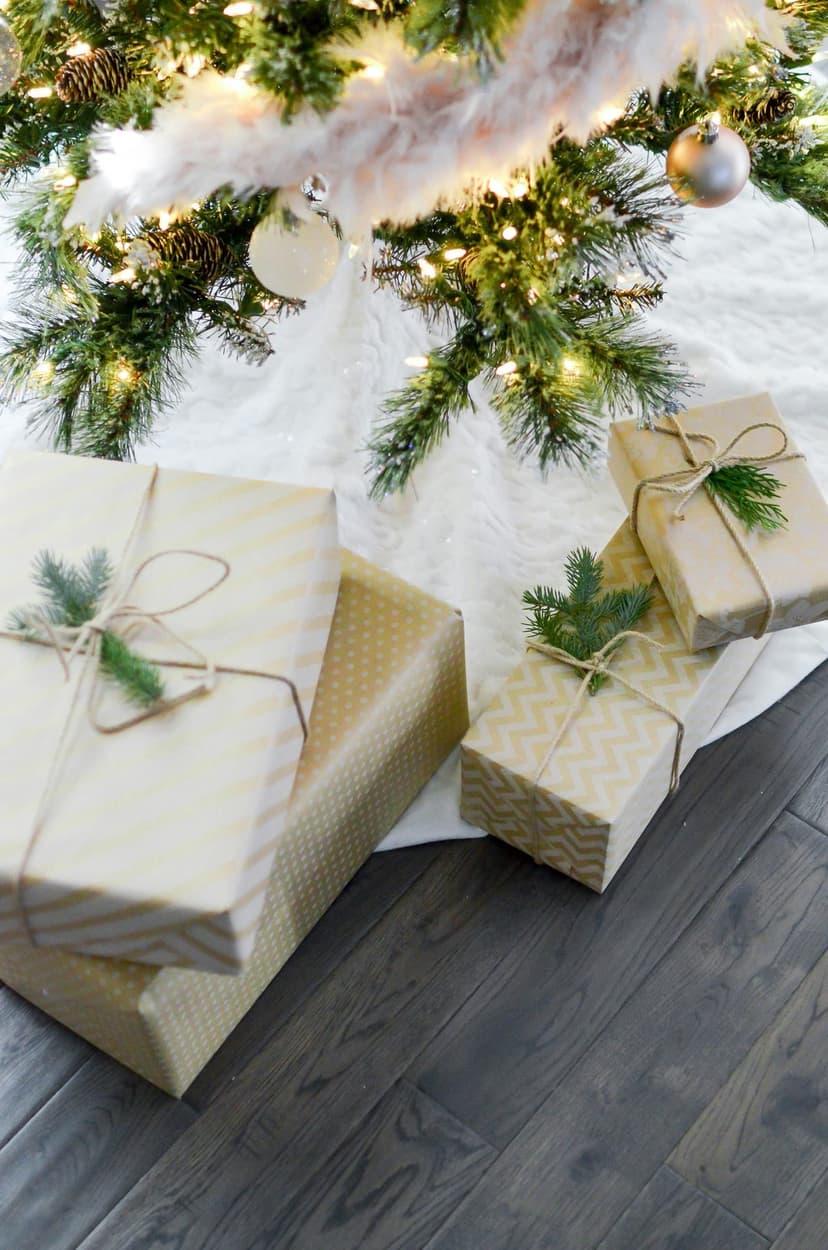 Christmas gifts in kraft and gold paper rest on white faux fur beneath decorated evergreen pine branches.