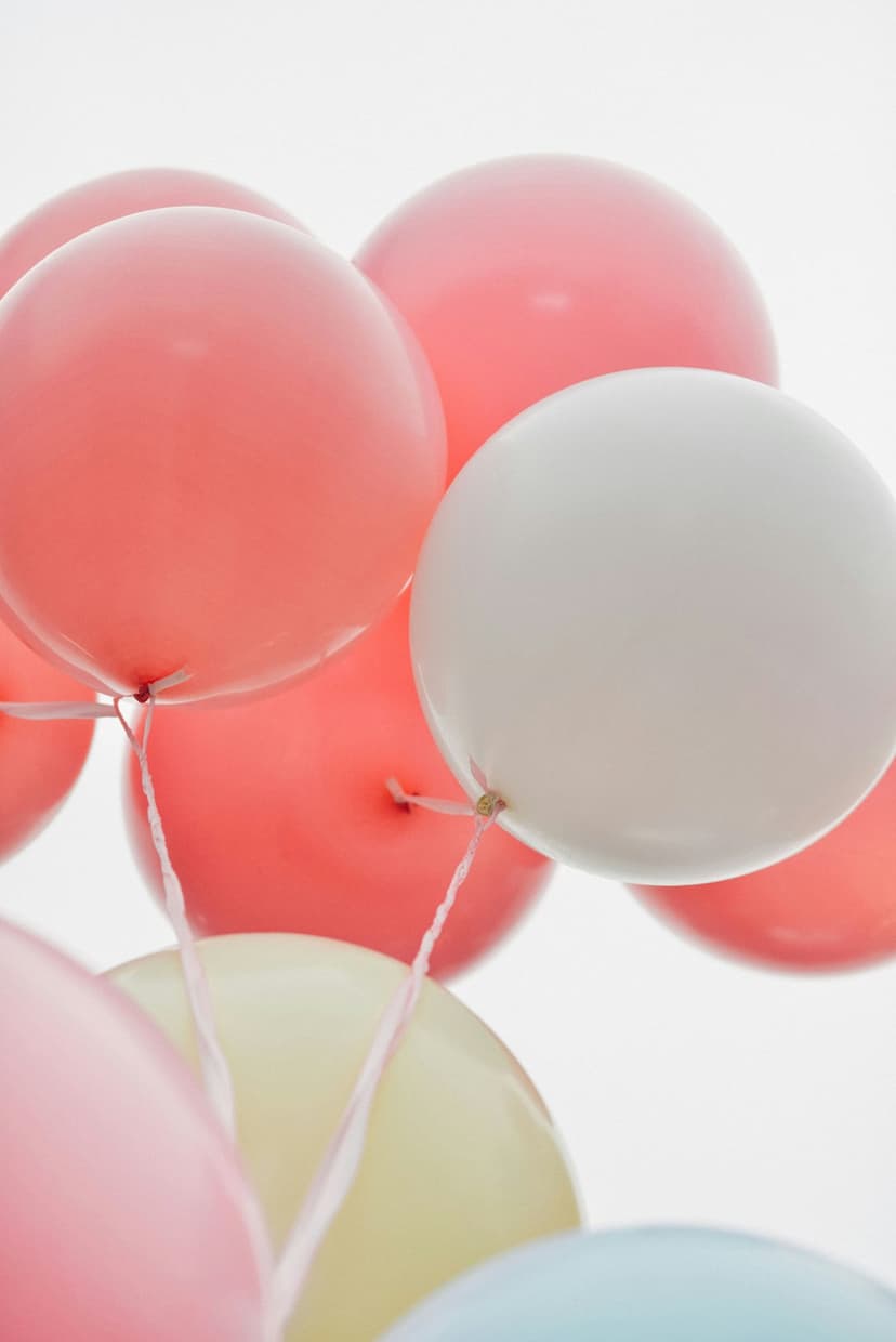 Pastel pink and yellow balloons float upwards against a clean white sky in a bright, minimalist composition.