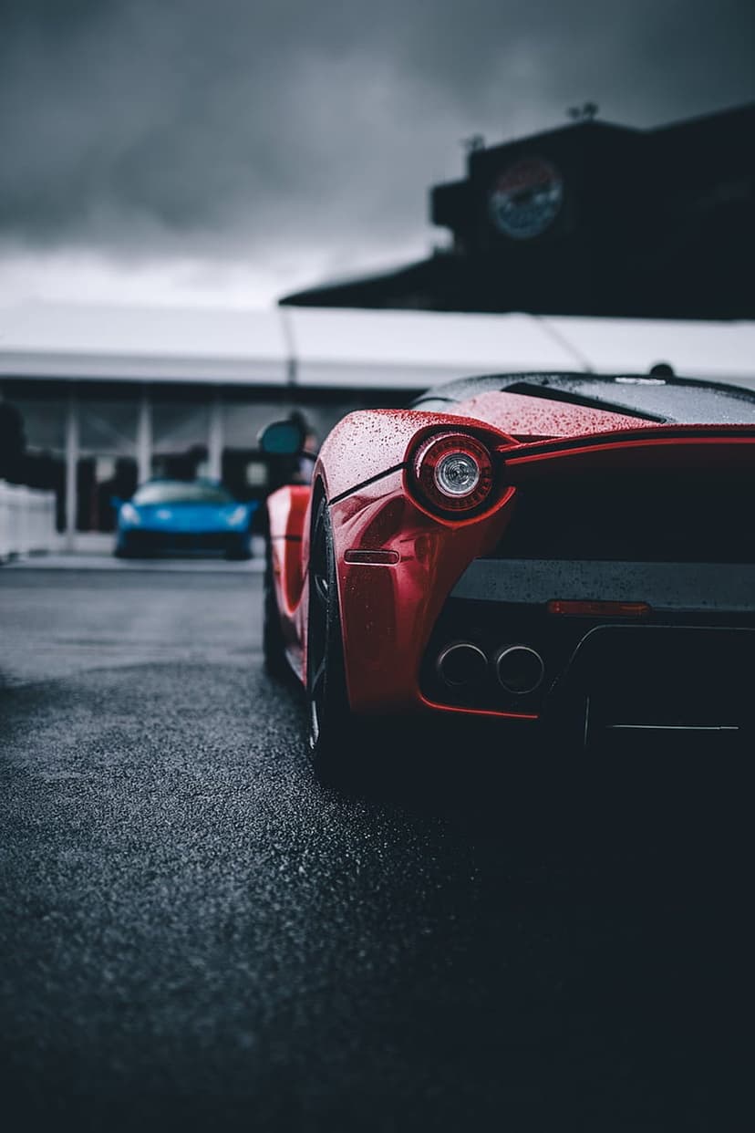 Red Ferrari rear bumper with raindrops and a blurred blue car parked on wet asphalt under a cloudy sky.