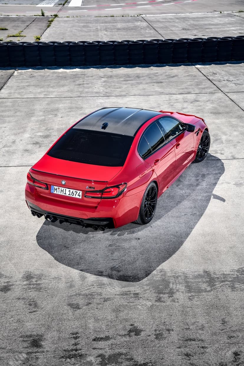 Red BMW M5 Competition sports sedan on a concrete track with a carbon fiber roof and black wheels.