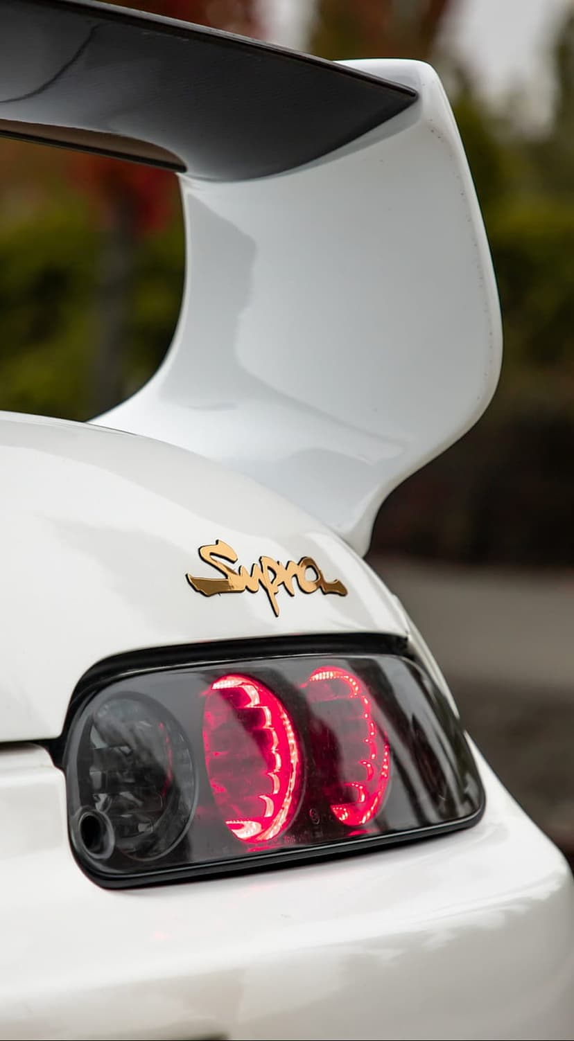 White Toyota Supra rear showing a large wing, red circular taillights, and a golden emblem on the bumper.