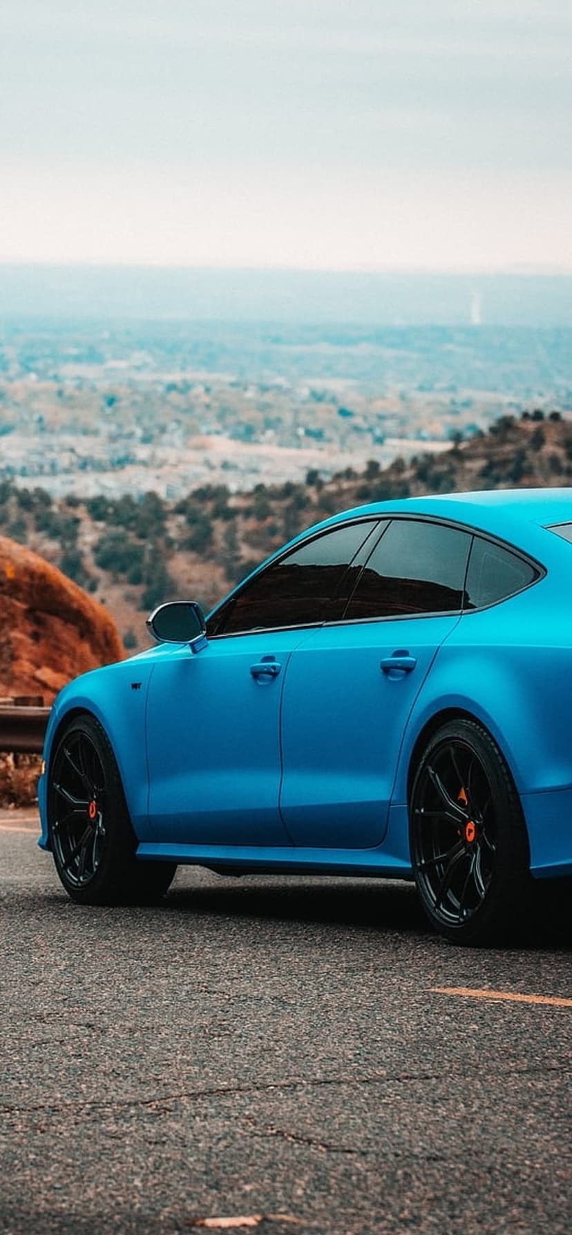 Blue Audi A7 sedan with black wheels parked on a winding mountain road overlooking a lush valley landscape.