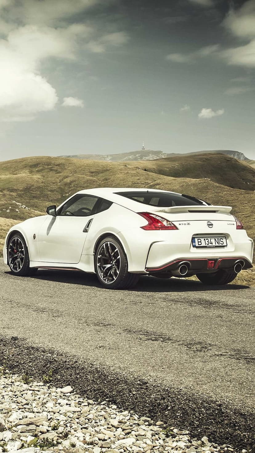 White Nissan 370Z sports car with red accents parked on a winding mountain road under a cloudy, dramatic sky.