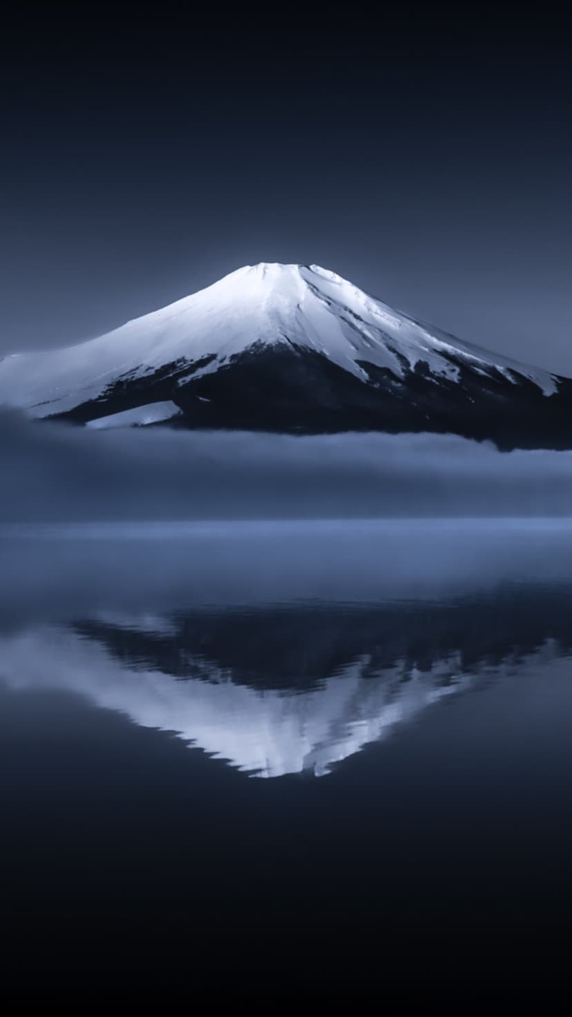 Mount Fuji reflected in dark water beneath a foggy sky with a snow-covered summit and deep monochromatic tones.