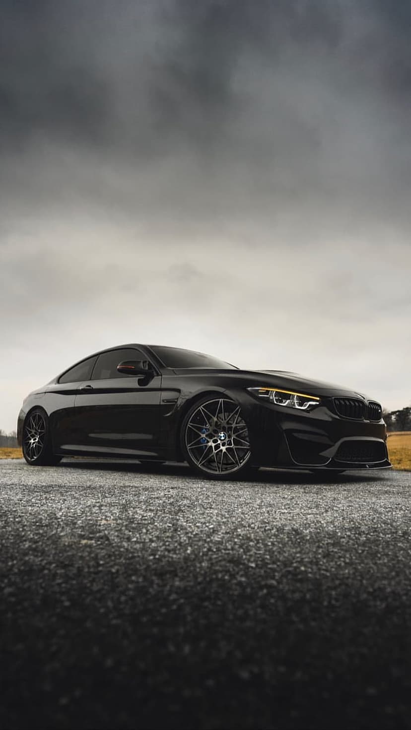 BMW M4 coupe in black paint parked on a textured road beneath a dramatic, grey cloudy sky at a low angle view.