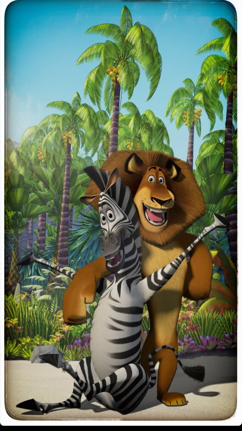 Alex the Lion and Marty the Zebra hugging amidst green palm trees and exotic plants under a bright blue sky.