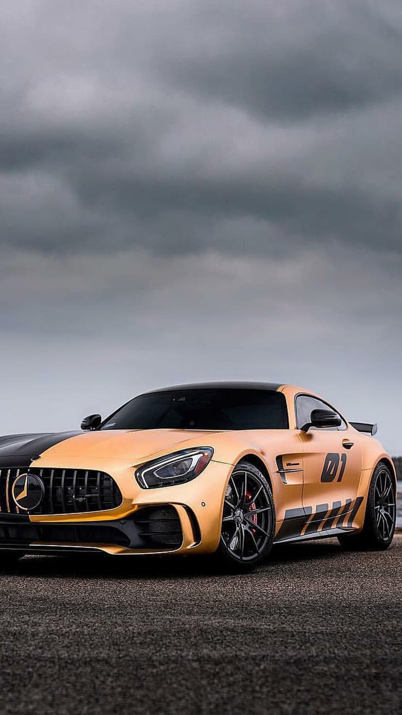 Golden Mercedes-AMG GT R with a black racing stripe and 01 side decal parked on dark asphalt under cloudy skies.