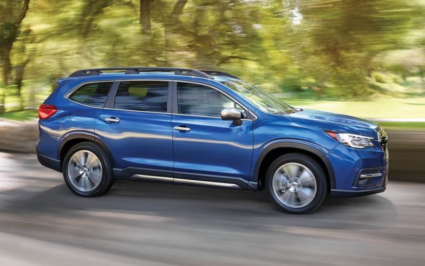 Blue Subaru Ascent SUV driving fast with motion blur on a sunlit road through a green forest background.