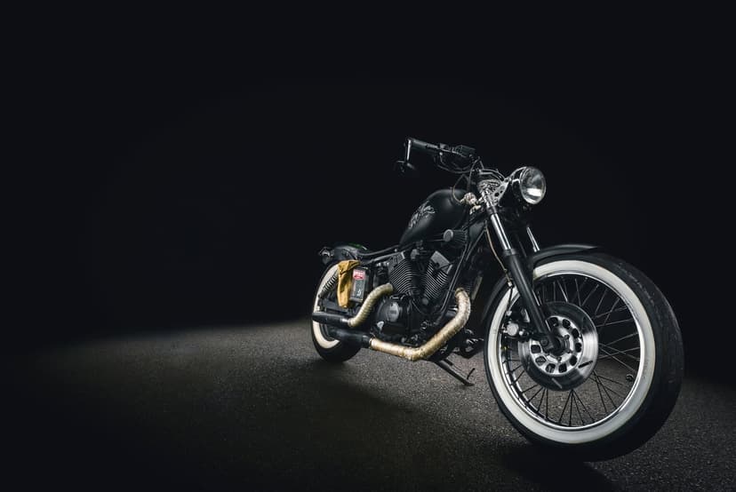Custom black cruiser motorcycle with white wall tires and gold exhaust headers against a dark studio backdrop.