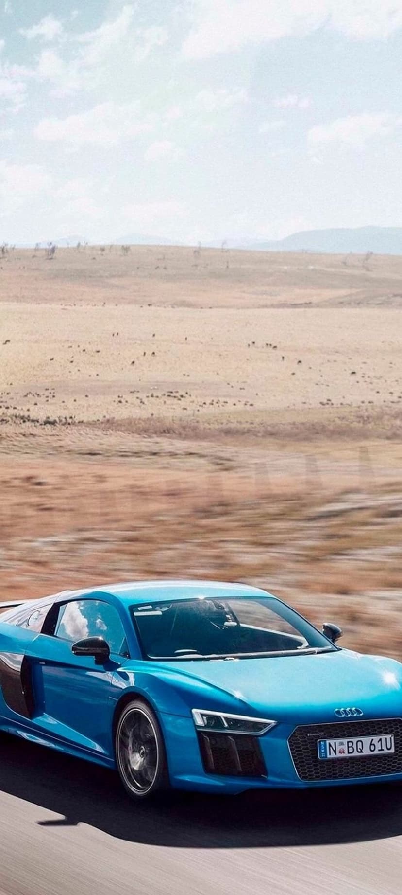 Blue Audi R8 sports car speeds through a dry, dusty desert terrain under a bright sky with rolling hills.
