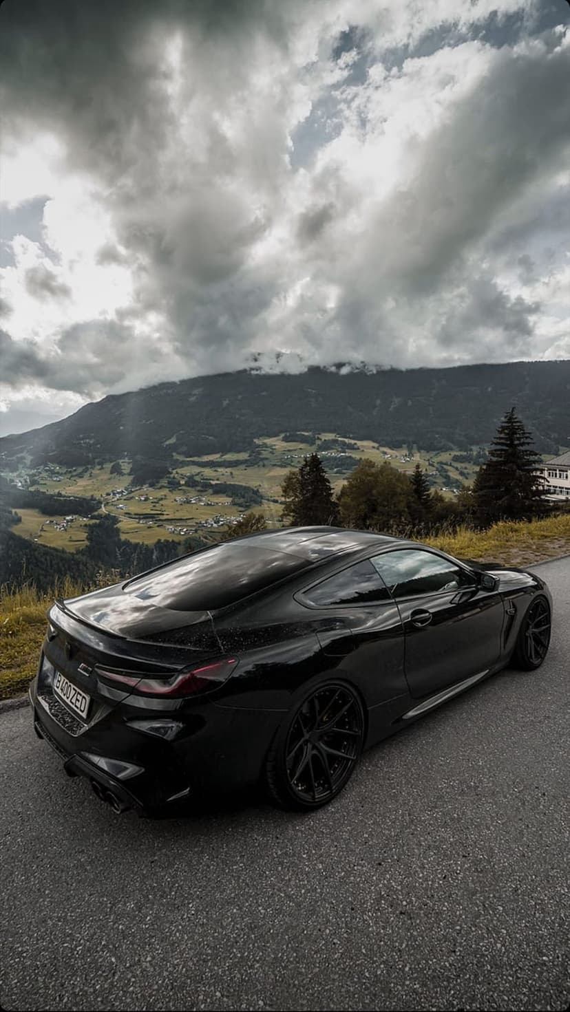Black BMW coupe parked on a paved mountain road under a dark, cloudy sky with jagged alpine peaks in the distance.