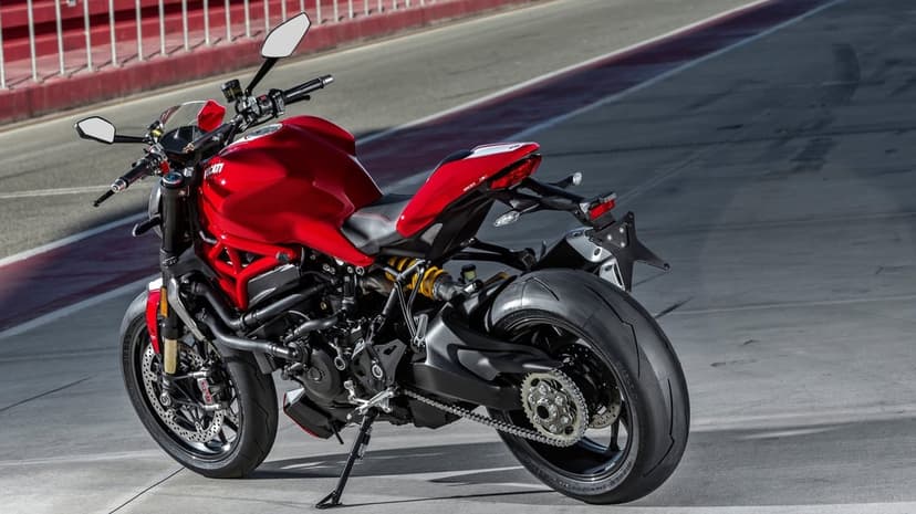 Red Ducati Monster sportbike on a grey racetrack with red and white curbs under bright natural daylight.