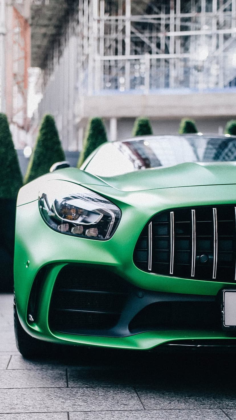 Matte green Mercedes-AMG GT front grille and headlights parked on stone tiles with blurred trees in the background.