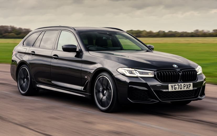 Black BMW 5 Series Touring M Sport driving on a road with motion blur against a green field and overcast sky.