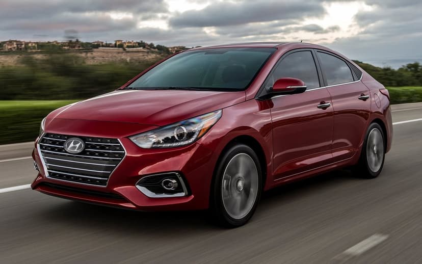 Red Hyundai Accent sedan driving on a highway under a cloudy sky with motion blur on the wheels and road.