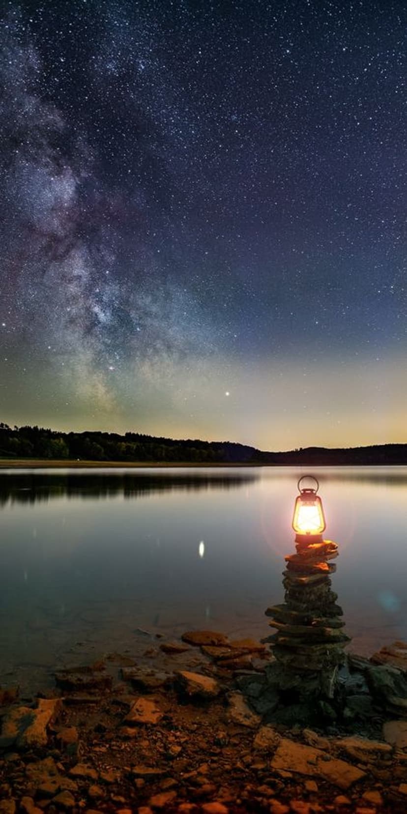 A small lantern atop stacked rocks glows warmly beside a calm lake reflecting the starry Milky Way galaxy above.