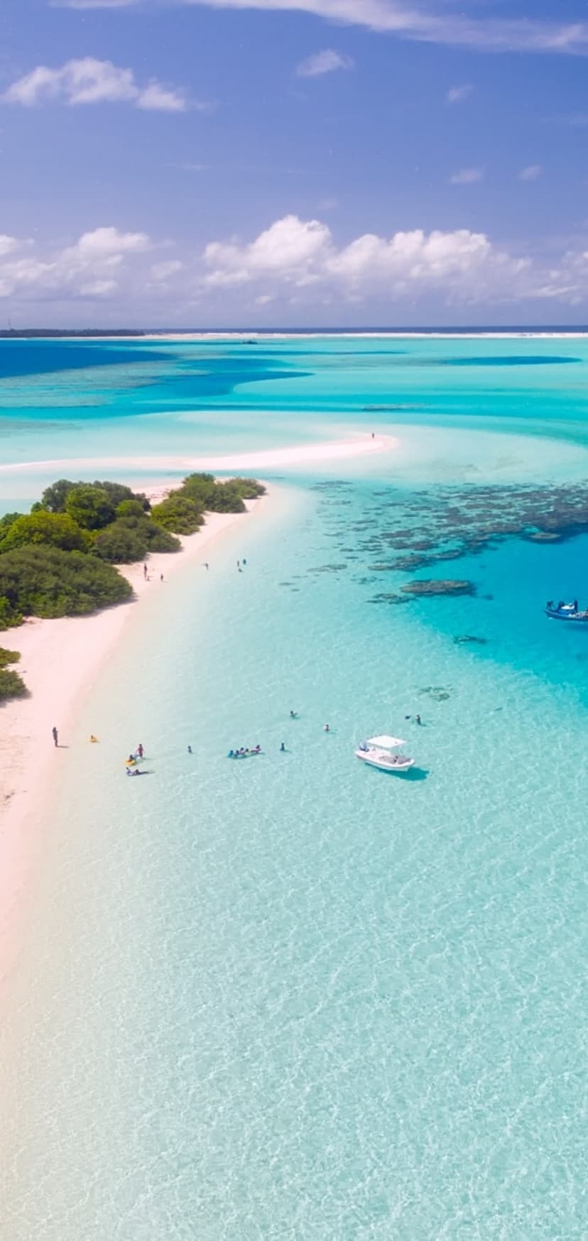 Curved ivory sandbar extending into transparent turquoise ocean waters with lush green palms and small boats.