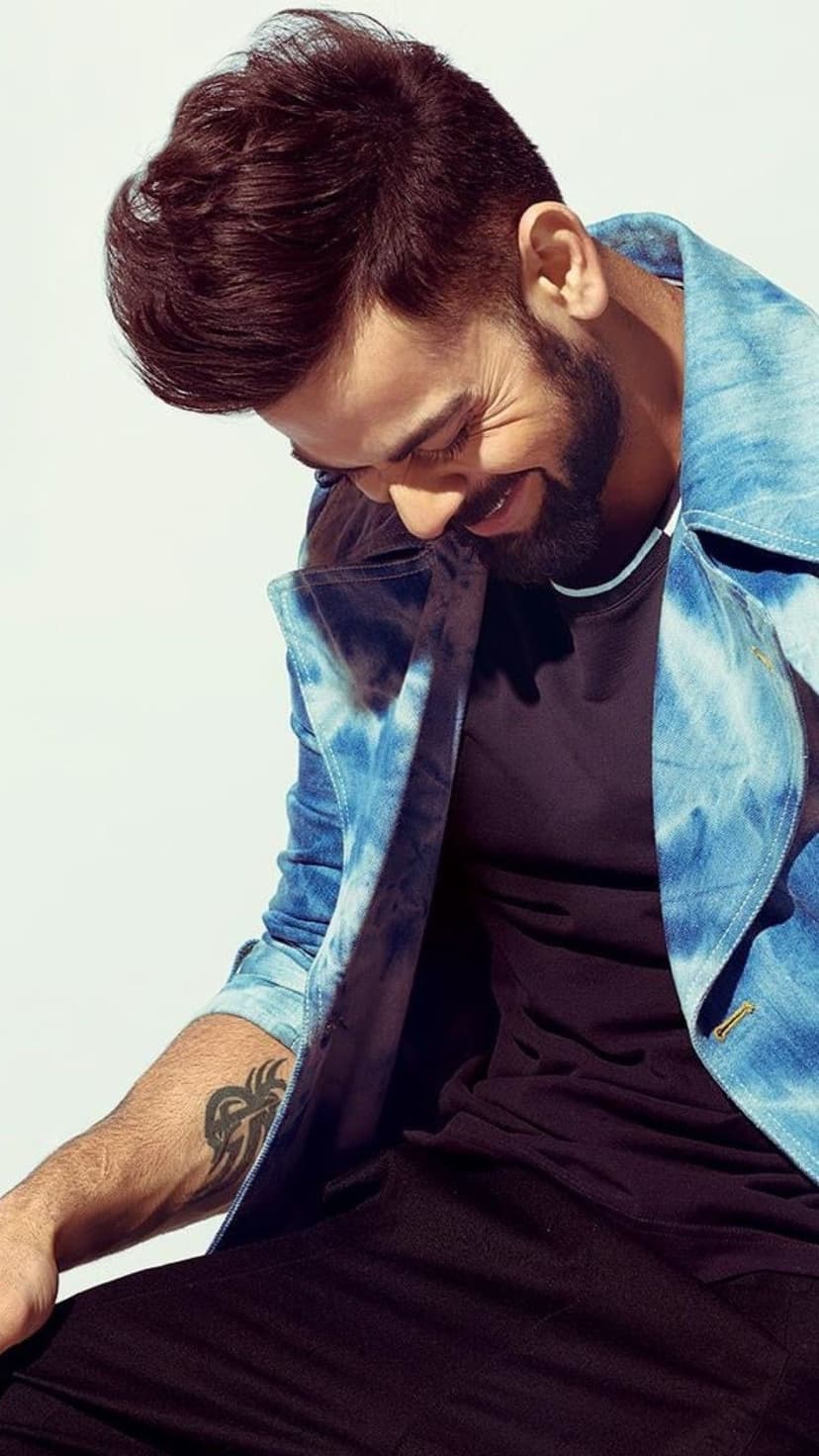 Virat Kohli laughs with head bowed wearing a tie-dye denim jacket and showing a detailed forearm tattoo.