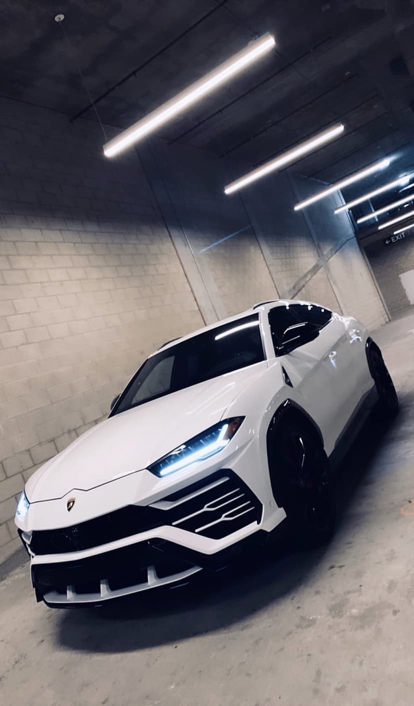 White Lamborghini Urus SUV with glowing headlights parked on concrete in a dimly lit modern parking garage.