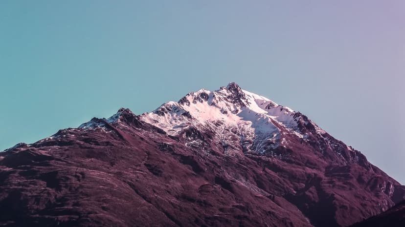 Snow-capped mountain peak towers against a teal and purple gradient sky with rugged dark rock faces in foreground.