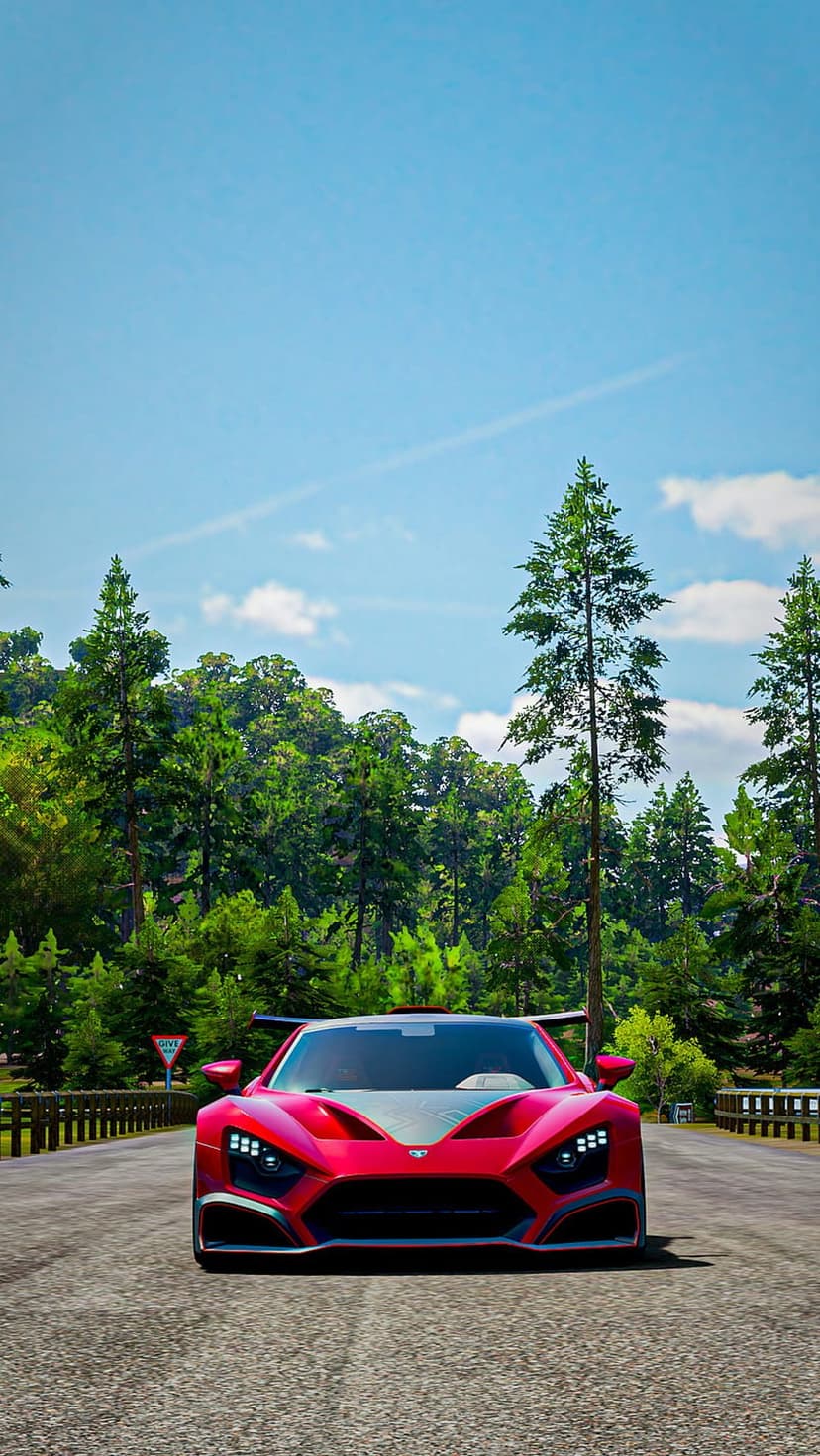 Red sports car driving head-on through a lush green forest under a clear blue sky on a paved mountain road.