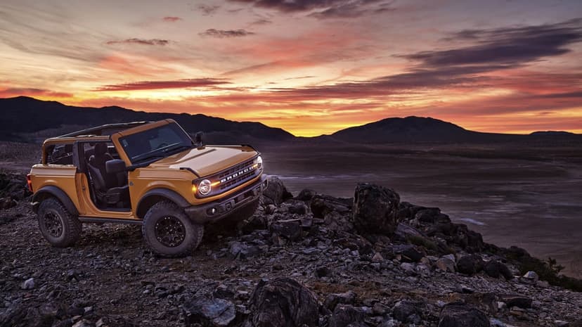 Yellow Ford Bronco parked on a rocky ridge under a vibrant orange and pink sunset sky in a desert landscape.