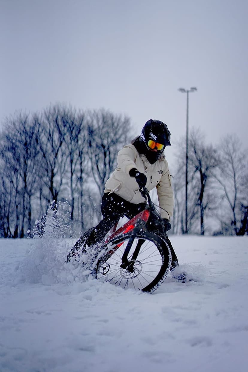 Cyclist on a fat tire bike kicks up a spray of white snow in a dense winter forest during a high-speed descent.