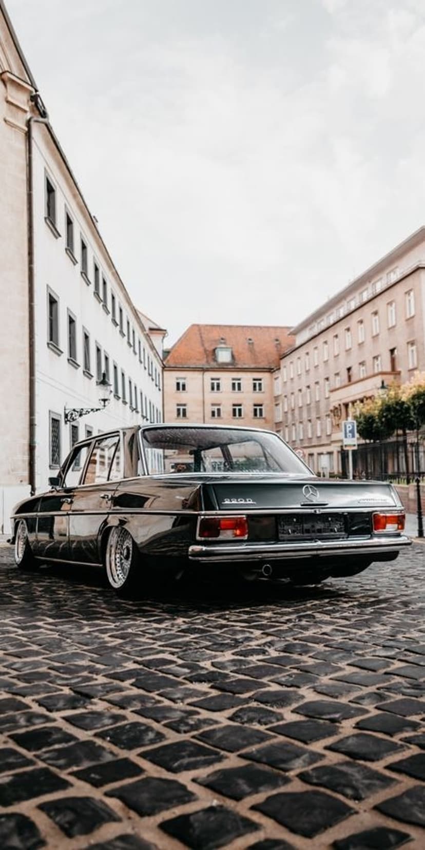 Dark Mercedes-Benz 280SE rear view on a cobblestone street with classic European architecture and chrome details.