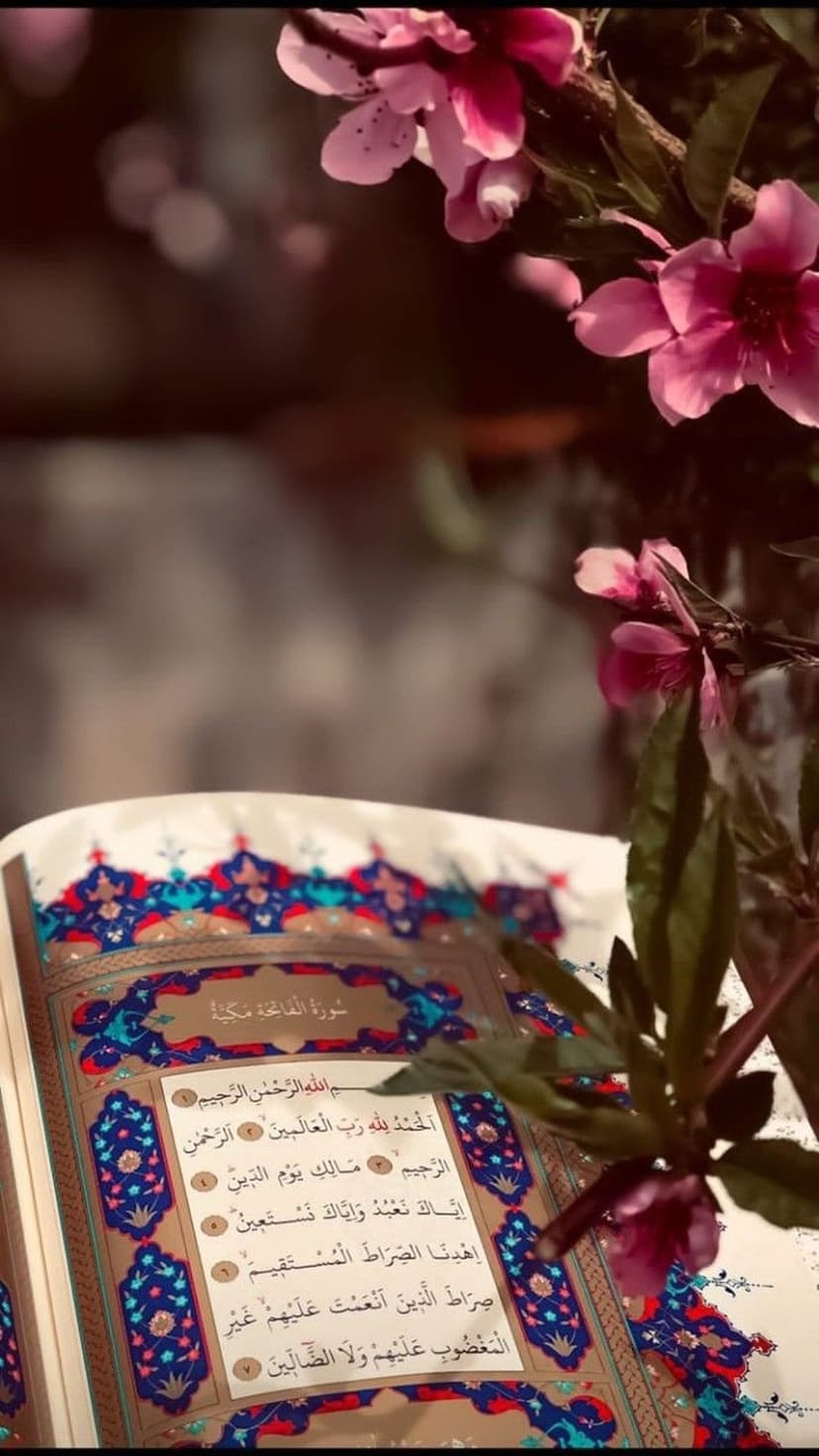 Open Quran with intricate Arabic calligraphy rests beside soft pink flowers in a bright, tranquil setting.