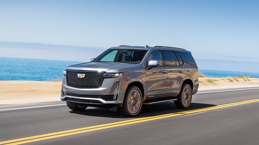 Silver Cadillac Escalade driving on a coastal road with blue ocean and clear sky in the background.