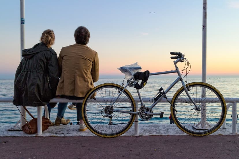 Two people sit on a bench with a bicycle nearby, overlooking a vibrant orange and pink sunset above calm sea water.