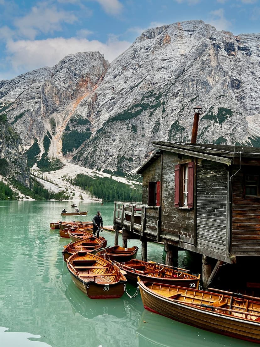 Rustic wooden boathouse and numbered rowing boats on a turquoise mountain lake with jagged peaks in the background.