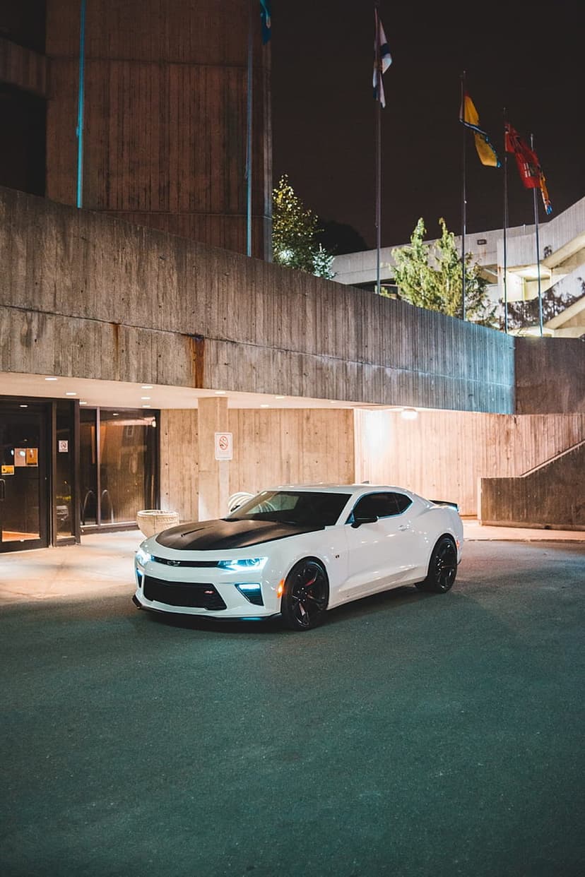 White Chevrolet Camaro with a black hood parked by a concrete brutalist building at night with headlights on.