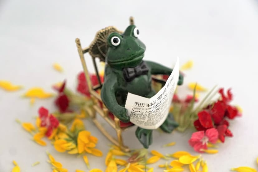 Green frog figurine wearing a bow tie sits in a rocking chair reading a miniature newspaper among flower petals.