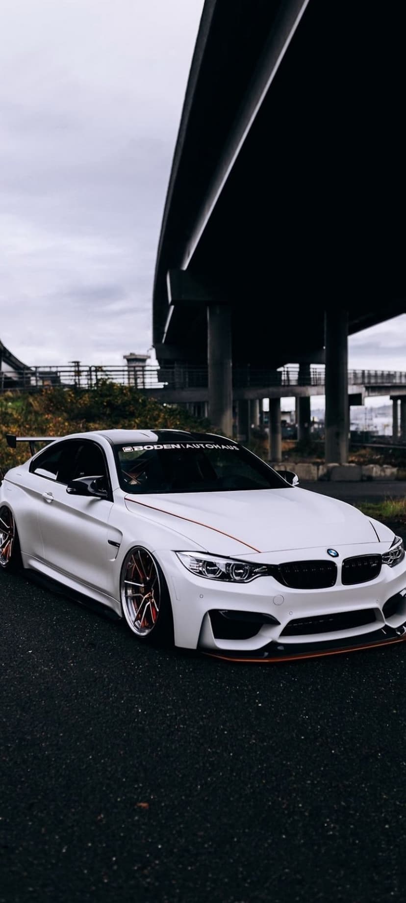 White BMW M4 coupe with a large rear spoiler and red-rimmed wheels parked on asphalt under a highway bridge.