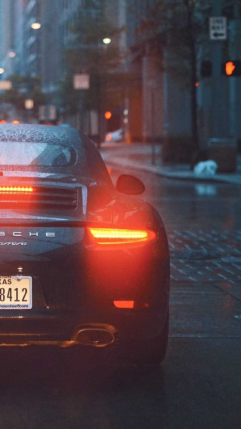 Porsche Carrera rear with glowing red taillights on a wet city street at night reflecting urban neon lights.