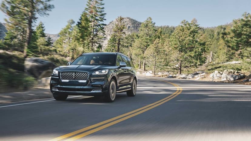 Dark blue Lincoln SUV driving along a winding asphalt road through a sunlit pine forest with mountains behind.
