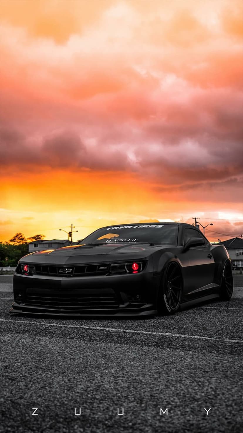 Matte black Chevrolet Camaro with glowing red headlights parked on asphalt under a dramatic orange sunset sky.