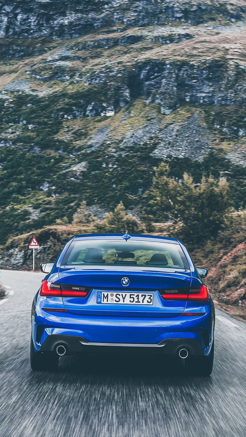 Blue BMW 3 Series sedan driving away on a winding mountain road with glowing red taillights and rocky cliffs.