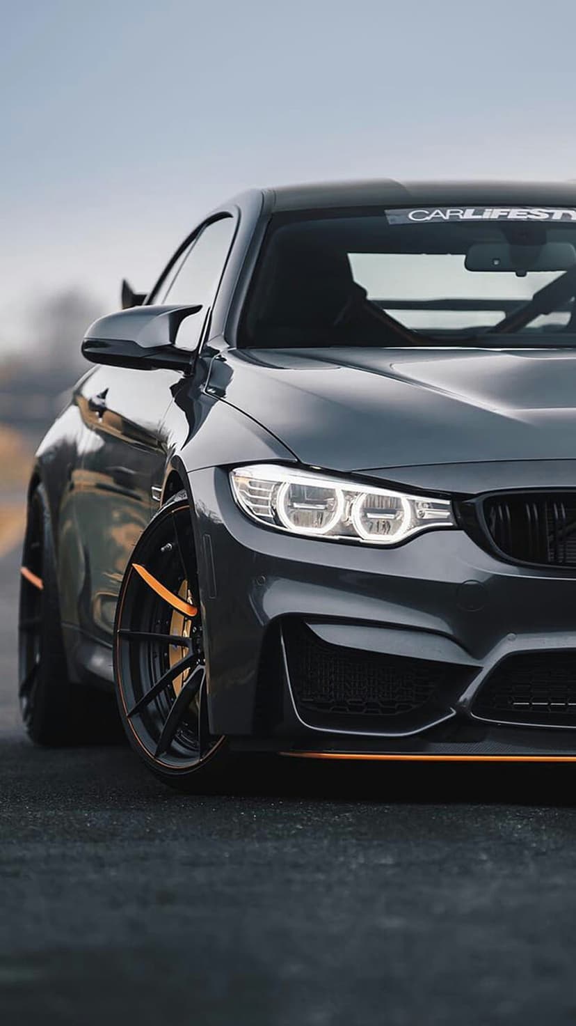 BMW M4 Coupe in dark grey with orange trim and illuminated LED headlights on an asphalt road at a low angle.