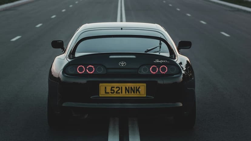 Black Toyota Supra with glowing circular taillights cruises on an empty road, captured from a rear angle.